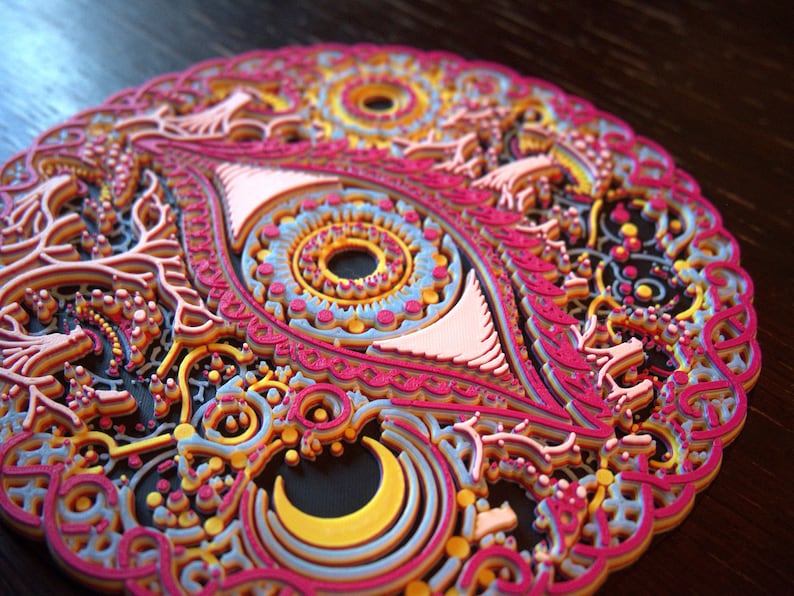 May include: A circular, multi-layered wall art piece featuring an intricate eye design at the center. The artwork is composed of stacked, laser-cut elements in vibrant shades of hot pink, bright yellow, light blue, and orange against a black base.