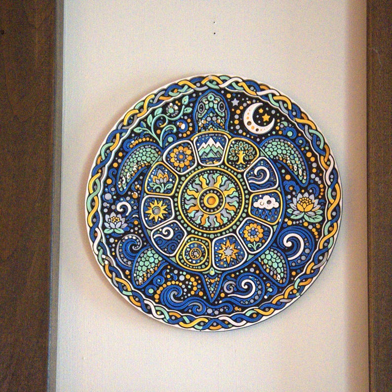 May include: A circular wall plaque featuring a detailed illustration of a sea turtle. The shell is divided into segments containing symbols like a sun, moon, mountains, and flowers. The design uses a blue, yellow, and black color palette.