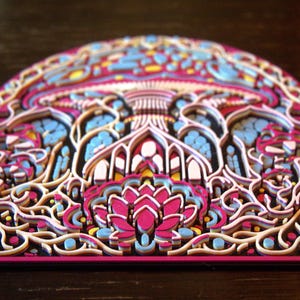 May include: A multi-layered, arched wall art piece featuring intricate, laser-cut designs in vibrant shades of magenta, light blue, yellow, and white. The composition displays a central lotus-like floral motif surrounded by swirling, organic patterns.