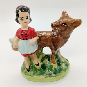 Może przedstawiać: Ceramiczna figurka przedstawiająca osobę z ciemnymi włosami w czerwonej koszuli w czarne kropki i jasnoniebieskim fartuchu, stojącą obok brązowego jelenia. Obie postacie znajdują się na zielonej podstawie z małym okrągłym otworem pośrodku.
