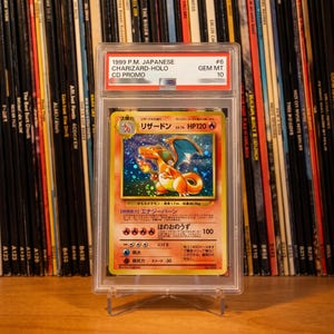 May include: A graded 1999 Japanese Charizard holographic trading card encased in a clear plastic protective slab. The card features a fire-breathing dragon illustration with a gold border, Japanese text, and a Gem Mint 10 label at the top.