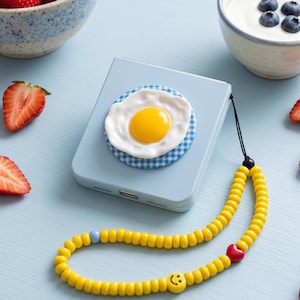 May include: A light blue square phone case featuring a raised fried egg decoration on a blue and white checkered circle. A yellow beaded phone strap is attached, accented with a single red heart bead and a small yellow smiley face bead.