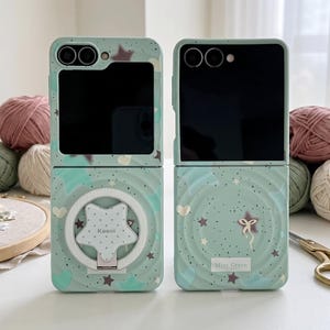 May include: Two mint green foldable phone cases with a speckled pattern featuring stars and hearts. The left case has a white star-shaped ring holder with the text Koooi, while the right case features a small bow graphic and the text Mint Green.