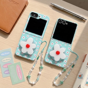 May include: Two light blue foldable phone cases featuring a white daisy pattern with red centres. Each case includes a large white flower-shaped grip and a beaded phone strap with small star-shaped beads attached to the side of the device.