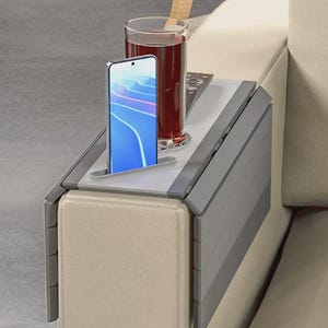 May include: A gray flexible sofa arm tray with a flat surface, a circular cutout for a glass, and a narrow slot for a smartphone. The tray features a segmented design that drapes over the armrest, holding a smartphone, a glass of dark liquid, and a remote control.