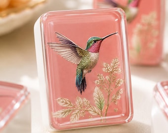 Könnte beinhalten: Ein rechteckiger Briefbeschwerer aus Glas mit rosafarbenem Hintergrund, der eine detaillierte Illustration eines Kolibris mit grünen, roten und blauen Federn zeigt, der über einem zarten, goldfarbenen Blattzweig mit kleinen weißen Blüten schwebt.