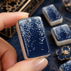 Peut inclure: Une tuile de mah-jong rectangulaire avec une surface bleu foncé brillante ornée d'un motif floral blanc qui s'estompe du haut vers le bas. La tuile possède une base épaisse et lisse de couleur blanc cassé avec des bords arrondis sur la face.