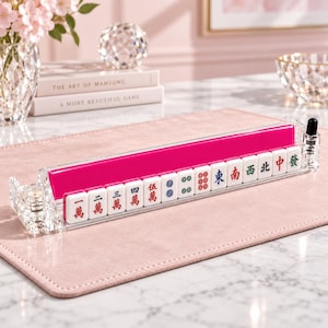 May include: A clear acrylic Mahjong tile rack featuring a vibrant hot pink back panel. The rack holds a row of white Mahjong tiles, each displaying distinct Chinese characters and symbols in red, blue, and green ink, supported by crystal-like ends.
