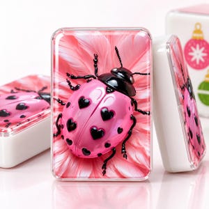 May include: A rectangular white container featuring a glossy pink ladybug with black heart-shaped spots resting on a background of soft pink feathers. The ladybug has a shiny black head and black legs, creating a high-contrast, vibrant visual display.