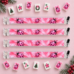 May include: Four clear acrylic mahjong tile racks featuring a pink background with repeating pink ladybug patterns. Each rack has a black end cap and a metal screw mechanism. Scattered around are various mahjong tiles with ladybug and holiday designs.