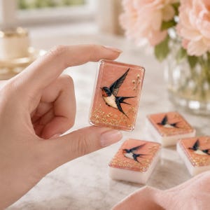 May include: A rectangular resin tile featuring a swallow bird illustration with a red throat, black wings, and white underbelly, set against a peach-colored background with gold leaf accents. The tile has a white base and a clear, glossy finish.