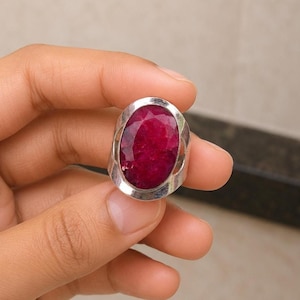May include: A silver ring featuring a large, oval-shaped, faceted deep pink gemstone set in a polished metal bezel. The band of the ring has a wide, open-work design with cut-out sections on the sides, showcasing a bold and substantial appearance.
