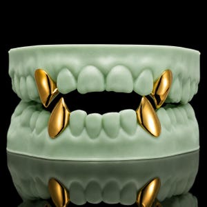 May include: A set of four polished gold tooth caps fitted onto a light green dental model. The gold caps are shaped like pointed fangs and are positioned on the upper and lower canine teeth of the dental mold, creating a vampire-like appearance.