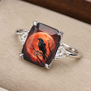 May include: A silver-toned ring featuring a large rectangular center stone with a printed image of a black crow perched on a branch against a large red moon. The center stone is flanked by two smaller triangular clear stones held by metal prongs.