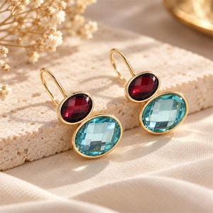 May include: A pair of gold-tone drop earrings featuring two oval-shaped faceted stones each. The top stone is a deep red, while the larger bottom stone is a bright light blue. The earrings are suspended from simple gold-tone wire hooks.
