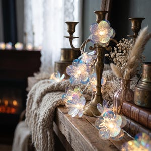 May include: A string of LED fairy lights featuring translucent, iridescent flower-shaped covers. Each flower has five petals with a glowing centre. The lights are draped around a brass candelabra, creating a soft, warm illumination.