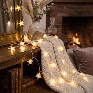 May include: A string of LED fairy lights featuring translucent star-shaped covers that emit a warm white glow. The lights are draped across a textured cream-colored knit throw, with the clear wire connecting each individual star-shaped bulb.