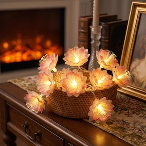 May include: A string of LED flower lights featuring soft pink and white petals with illuminated fiber optic centers, arranged inside a round woven rattan basket. The lights emit a warm glow, highlighting the layered petal structure.