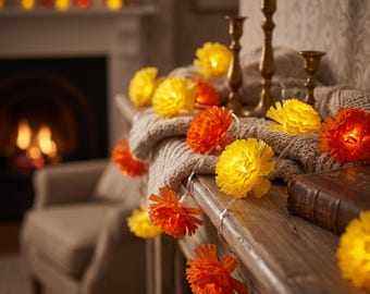 May include: A string of decorative lights featuring yellow and orange flower-shaped covers. The illuminated floral bulbs are arranged along a thin wire, resting on a wooden surface alongside a textured beige knit fabric and brass candle holders.