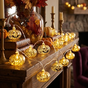 May include: A string of decorative LED lights featuring small, gold-toned glass pumpkin-shaped bulbs with carved jack-o'-lantern faces. The lights emit a warm glow and are connected by a thin, clear wire, arranged across a wooden mantelpiece.