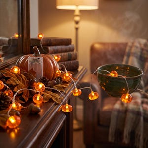 May include: A string of orange pumpkin-shaped LED lights with a clear wire. The small, glowing pumpkin bulbs are spaced along the cord, which is draped over a surface with autumn-themed decorations including a large pumpkin and leaves.