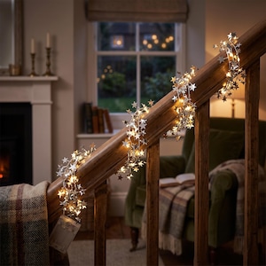 May include: A string of silver star-shaped fairy lights wrapped around a wooden staircase banister. The lights emit a warm glow, illuminating the metallic star cutouts. A clear plastic battery pack hangs from the bottom of the light strand.