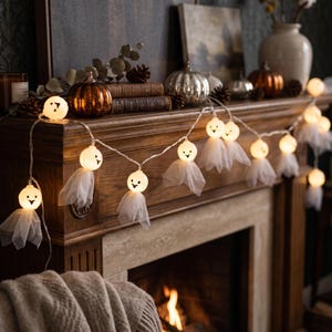 May include: A string of decorative LED lights featuring small white ghost figures. Each ghost consists of a round, illuminated head with a printed black face and a sheer, white fabric skirt draped underneath, connected by a thin, clear wire.