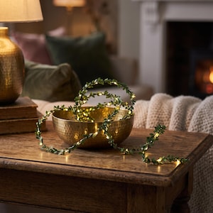 Può includere: Una catena luminosa con foglie verdi avvolta all'interno di una ciotola rotonda in metallo dorato martellato. La ghirlanda decorativa presenta piccole foglie verdi artificiali intervallate da minuscole luci LED dai toni caldi e soffusi.