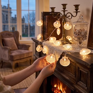 May include: A string of LED fairy lights featuring translucent, shell-shaped covers. The lights emit a warm glow and are draped across a wooden mantelpiece, with one shell light held in focus. The design includes multiple individual seashell-shaped bulbs.