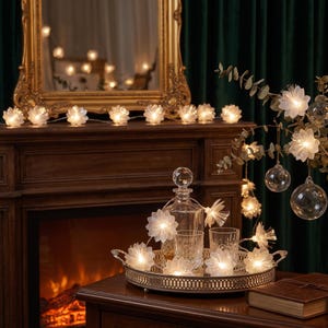 May include: A string of LED fairy lights featuring translucent white flower-shaped covers. The lights are arranged around a clear glass decanter and two matching drinking glasses on a metallic tray, with additional lights draped on a branch.