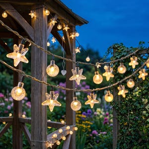 May include: A string of outdoor decorative lights featuring alternating clear glass globes and translucent star-shaped bulbs. The lights are strung along a wooden structure, emitting a warm glow against a twilight sky, with a small white heart ornament.