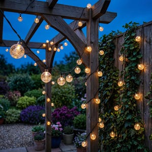 May include: A string of globe-shaped LED lights with a textured, bubble-like interior pattern, draped across a wooden pergola structure. The warm, glowing lights are suspended on a dark wire, illuminating the surrounding outdoor garden area.