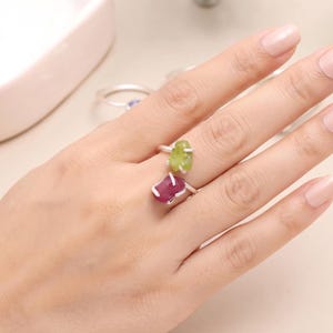 Könnte beinhalten: Ein Silberring mit zwei rohen, ungeschliffenen Edelsteinen, die von Metallkrallen gehalten werden. Ein Stein ist in einem leuchtenden Limettengrün, der andere in einem tiefen Magentarot. Der Ring besteht aus einem dünnen, metallischen Band.