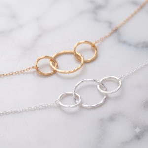May include: Two delicate necklaces, one in gold and one in silver, each featuring three interconnected, hammered-texture circular rings as a central pendant. The chains are thin and metallic, showcasing a simple design with a reflective finish.