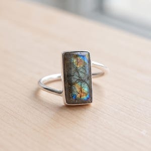 May include: A silver ring featuring a rectangular labradorite gemstone. The stone displays a grey base color with iridescent flashes of blue and gold. The setting is a simple metal bezel, and the band is a thin, smooth, polished silver wire.