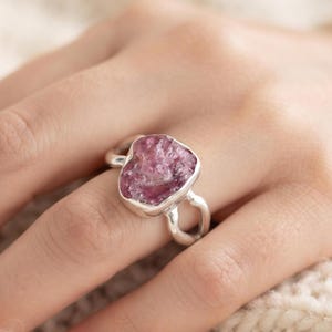 Könnte beinhalten: Ein Silberring mit einem rohen, ungeschliffenen rosa Edelstein in einer polierten Metallfassung. Die Ringschiene teilt sich in zwei glatte, getrennte Silberbögen, die zur zentralen Fassung führen und die natürliche, raue Textur des Steins betonen.