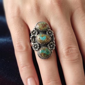 May include: A sterling silver ring featuring three oval-shaped turquoise stones with brown matrix patterns. The stones are arranged vertically and surrounded by a twisted rope border and four small silver flower accents on the sides of the band.