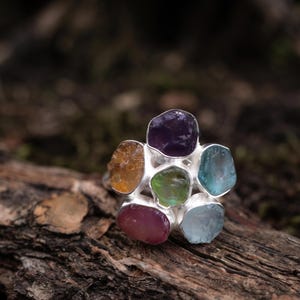Könnte beinhalten: Ein Silberring mit einer Anordnung aus sechs rohen, ungeschliffenen Edelsteinen in Blütenform. Die Steine umfassen einen tiefvioletten Amethyst, einen goldenen Citrin, einen hellgrünen Peridot, einen dunkelrosa Rubin und zwei hellblaue Aquamarine.