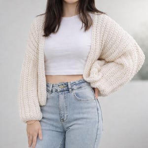 May include: Cream-colored, chunky knit cardigan with long, voluminous sleeves. The open-front sweater is paired with a white crop top and light wash blue jeans. The cardigan's texture and relaxed fit create a cozy, casual aesthetic.