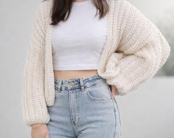 Könnte beinhalten: Cremefarbene, grob gestrickte Strickjacke mit langen, voluminösen Ärmeln. Der offene Pullover wird mit einem weißen Crop-Top und hellblauen Jeans kombiniert. Die Textur und die lockere Passform der Strickjacke schaffen eine gemütliche, lässige Ästhetik.