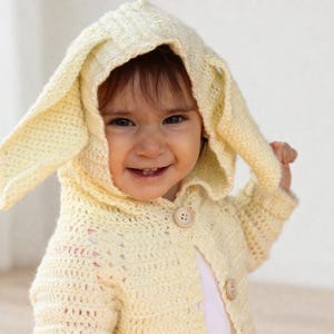 May include: A light yellow crochet hooded jacket with bunny ears. The jacket has two wooden buttons and is worn by a child. The hood features long, pointed ears, creating a playful, whimsical look. The crochet stitch is visible.