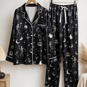 Könnte beinhalten: Ein schwarzer, langärmeliger Schlafanzug bestehend aus einem geknöpften Oberteil und einer Hose mit elastischem Bund und weißem Kordelzug. Das Set ist mit einem durchgehenden Muster aus Skeletten, Kürbissen, Geistern, Spinnennetzen und Fläschchen bedruckt.
