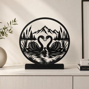 Könnte beinhalten: Eine kreisförmige Skulptur aus schwarzem Metall mit der Silhouette von zwei erwachsenen Schwänen und einem Schwanenküken auf dem Wasser. Das Design zeigt Schilf, Tannen und ein Gebirge im Hintergrund, montiert auf einem rechteckigen schwarzen Sockel.