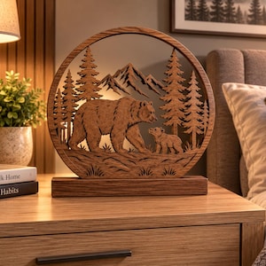 Könnte beinhalten: Ein kreisförmiges dekoratives Holzobjekt mit einem lasergeschnittenen Design eines großen Bären und eines Jungtiers, die durch einen Wald mit Bergen im Hintergrund laufen. Das Kunstwerk ist auf einem rechteckigen Holzsockel montiert und braun.