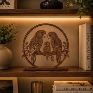 Könnte beinhalten: Ein kreisförmiges Wandkunstobjekt aus Holz mit einem lasergeschnittenen Design von zwei erwachsenen Papageien und einem kleinen Küken auf einem Ast. Die braune Holzskulptur steht auf einem rechteckigen Sockel und zeigt feine Details.