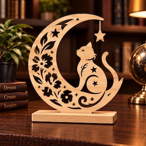 May include: A light-coloured wooden decorative ornament featuring a crescent moon shape with intricate floral cutouts and a cat silhouette. A small star hangs from the top of the moon. The entire piece is mounted on a rectangular wooden base.