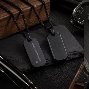 May include: Two black metal dog tag pendants on matching black box chains. One pendant features the engraved text "Love you forever" followed by the letter "D". The pendants have a brushed matte finish and are rectangular with rounded corners.