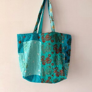 May include: A tote bag constructed from patchwork fabric in various shades of turquoise and teal. The bag features intricate embroidery with red and gold floral patterns, accented by white running stitches throughout the quilted surface.