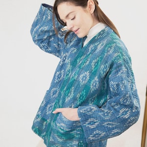 May include: A quilted jacket in shades of blue and teal featuring a repeating ikat-style pattern. The jacket has a V-neckline, long sleeves, and two front patch pockets. The fabric has a visible stitched texture throughout the entire garment.