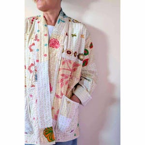Puede incluir: Una chaqueta de manga larga con un dise&ntilde;o de patchwork en tonos crema, rosa y verde con motivos florales. La prenda incluye un cierre frontal con botones de colores, dos bolsillos delanteros y costuras blancas visibles en la tela.